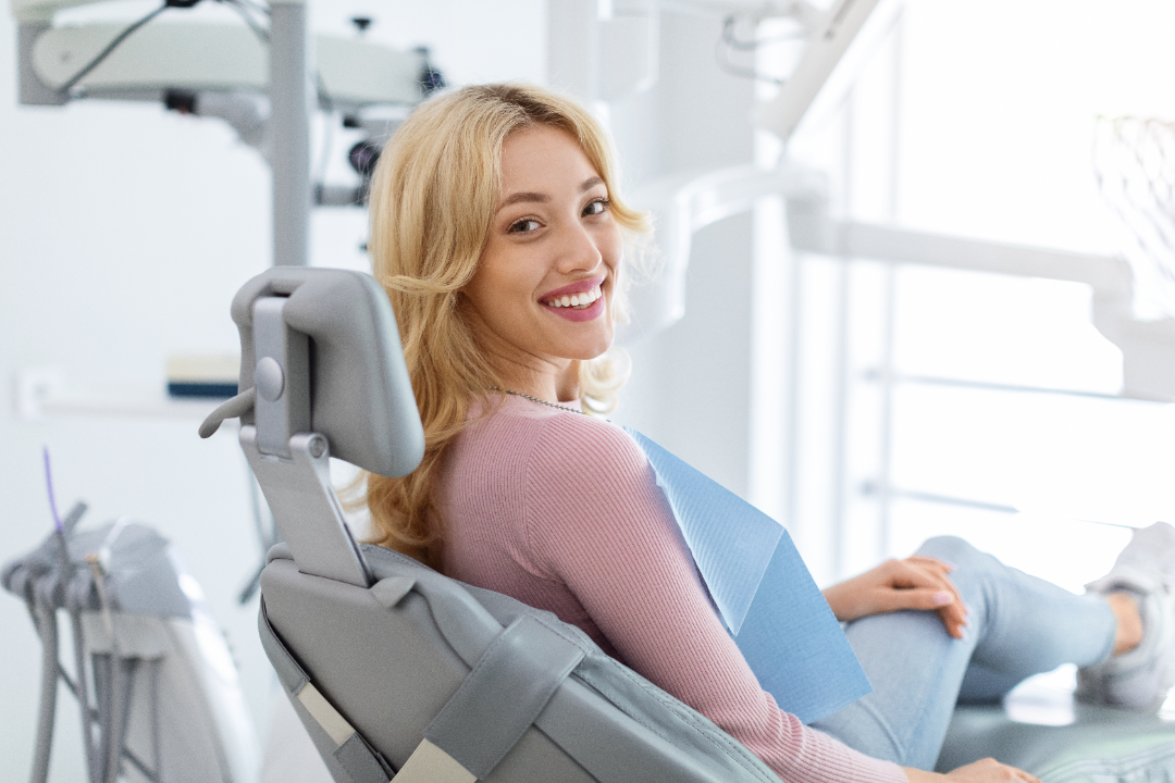 Smiling woman with blonde hair sitting in a dental chair wearing a pink sweater and blue dental bib.