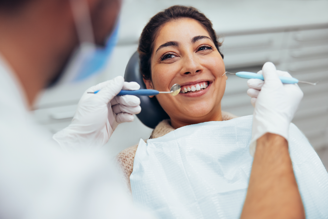 Smiling woman receiving a dental checkup from a dentist wearing gloves and holding dental tools.