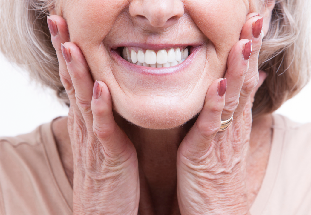 Close-up of an elderly woman smiling, with hands gently holding her face and showing white teeth and painted nails.