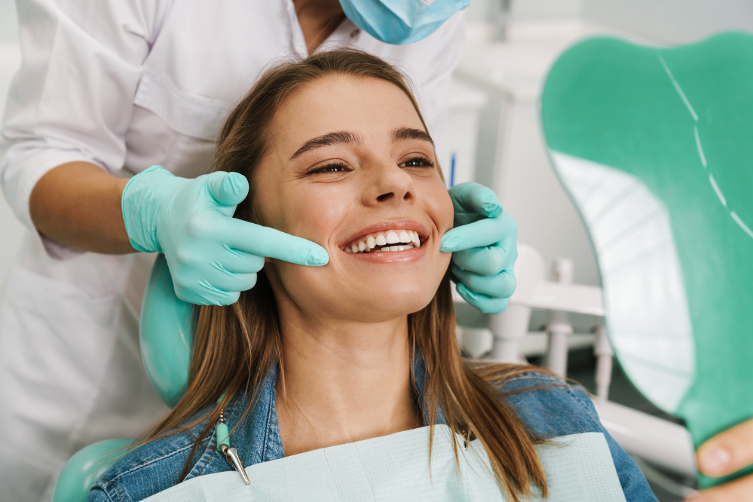 Dentist wearing gloves and mask pointing to a smiling woman's teeth as she looks into a mirror in a dental clinic.