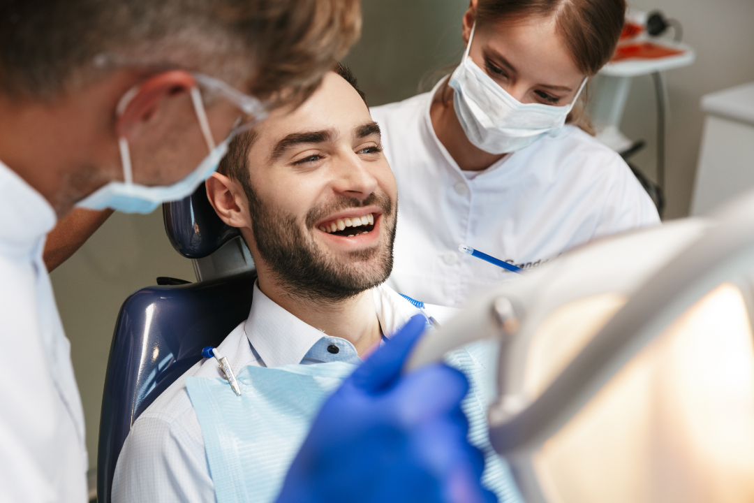 Smiling man sitting in dental chair during checkup with a dentist and dental assistant wearing masks.