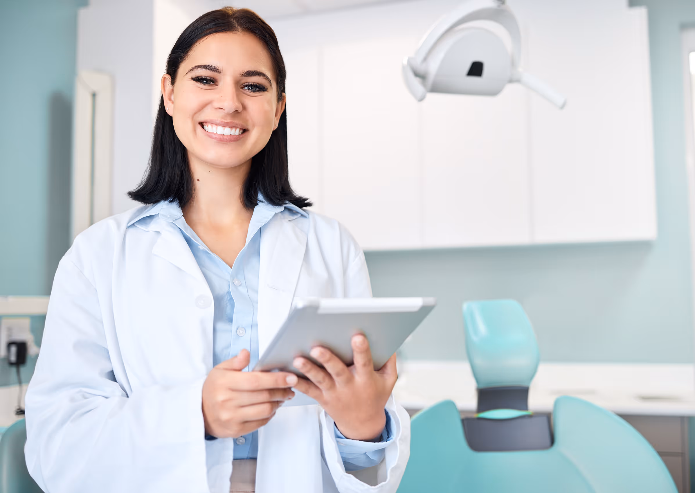 Smiling female dentist holding a tablet in a dental office with a dental chair and equipment in the background.