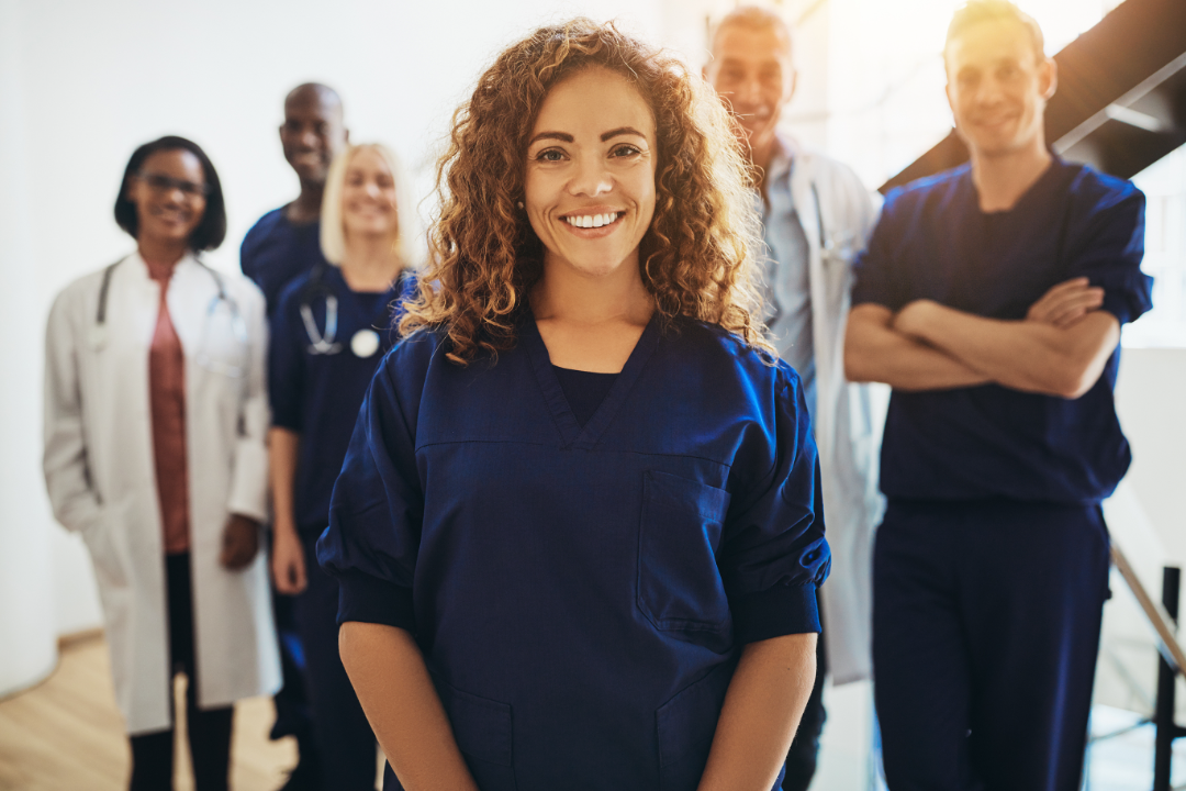 Smiling medical professional in blue scrubs standing confidently in front of a diverse healthcare team.